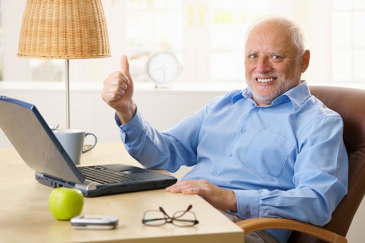 an older man sitting at a desk with his laptop and giving the thumbs up sign