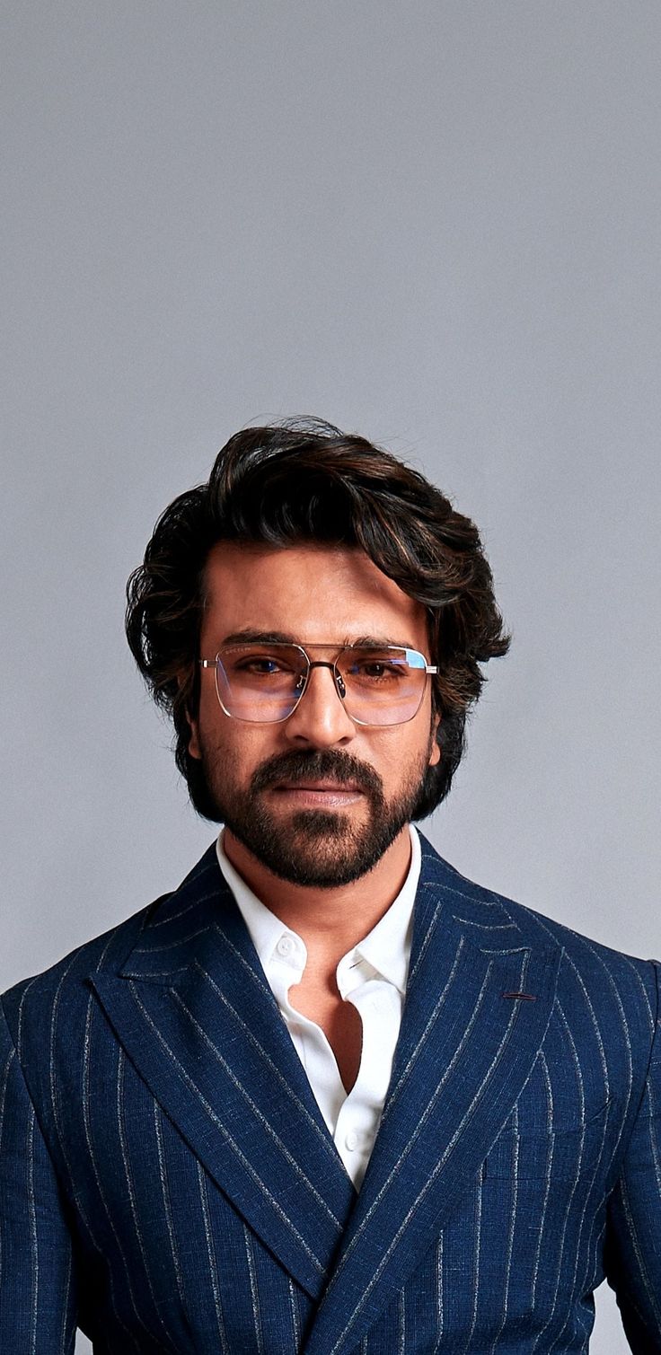 a man with glasses and a beard in a blue suit poses for a portrait against a gray background