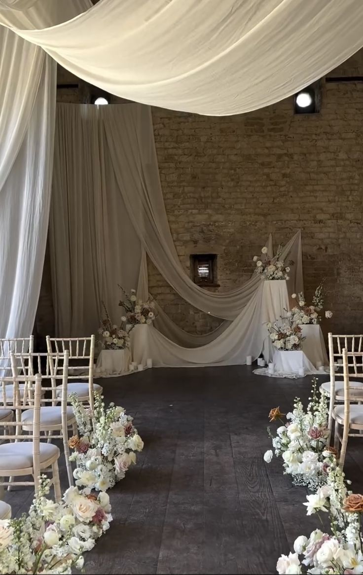 an indoor wedding venue with white draping and floral arrangements on the floor, along with chairs