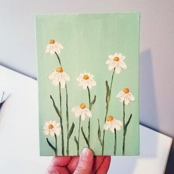 a hand holding up a small painting with daisies on the front and back of it