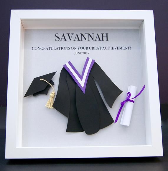 an origami graduation cap and gown is displayed in a white shadow box with purple ribbon