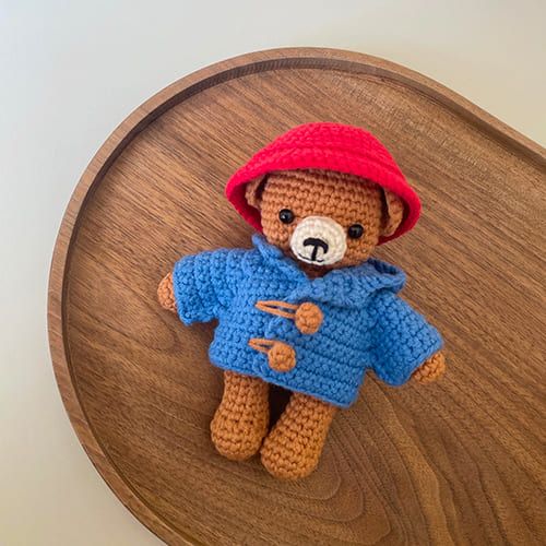 a crocheted teddy bear wearing a red hat and blue shirt on top of a wooden table