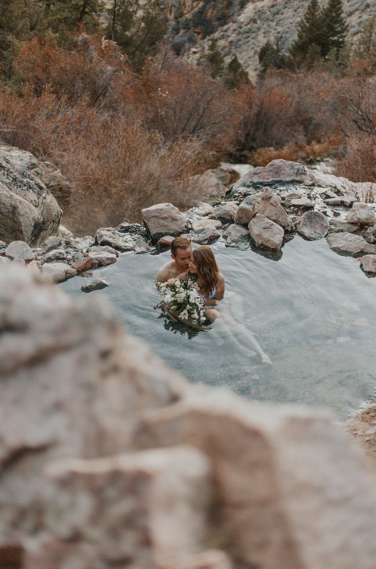 a man and woman are sitting in a hot tub surrounded by rocks, water and trees