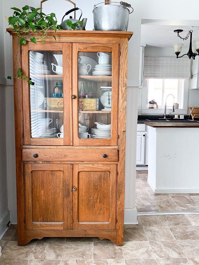 a wooden china cabinet with glass doors and plants on top, in the middle of a kitchen