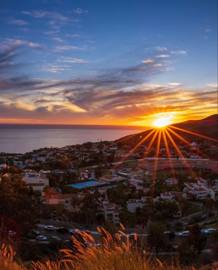 the sun is setting over a small town by the ocean with mountains in the background