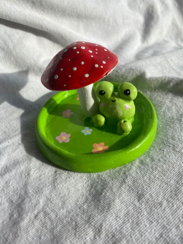 a small green frog sitting in front of a red mushroom on top of a white sheet