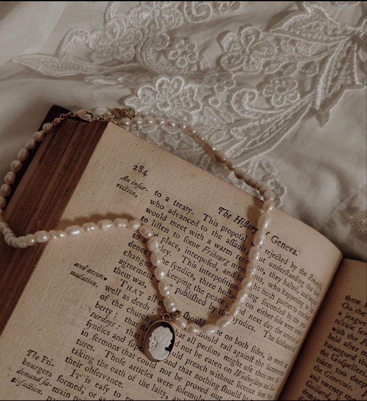 an open book with a necklace on it sitting on a bed next to a lace covered pillow