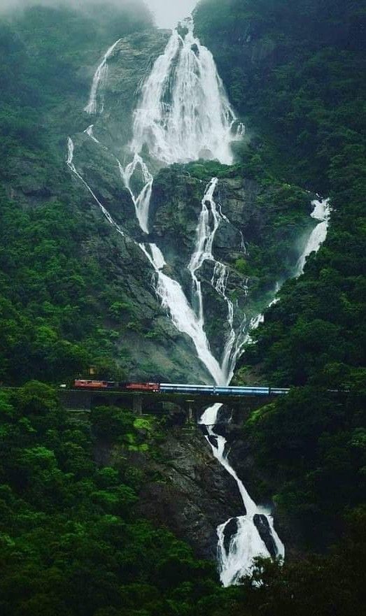 a train traveling over a bridge next to a lush green forest covered mountain side under a tall waterfall