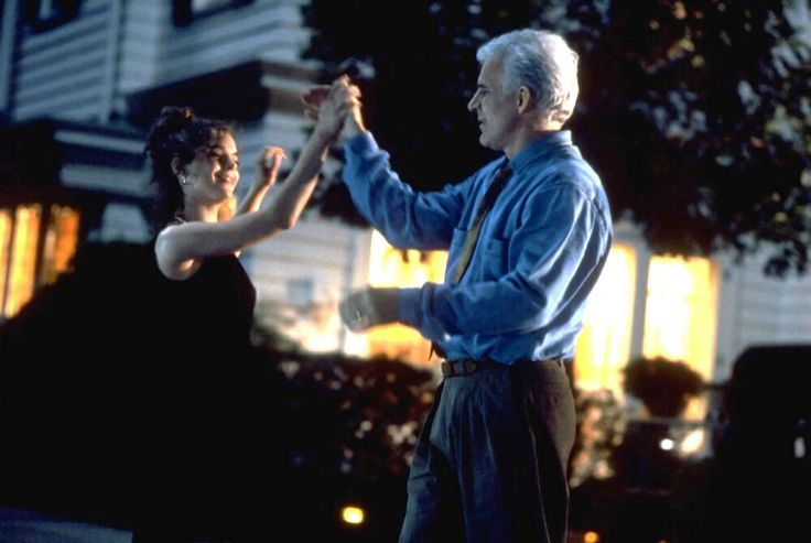 an older man and young woman dancing in front of a house at night with lights on