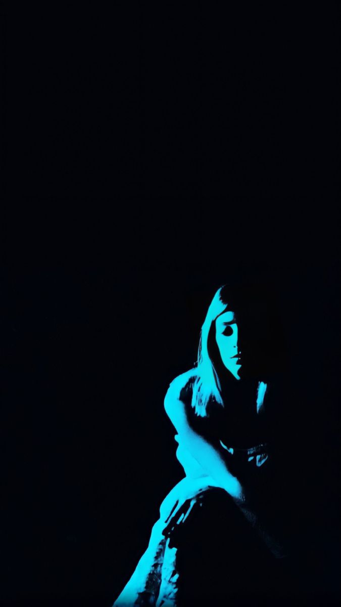 a woman is sitting in the dark with her arms around her neck and looking off to the side