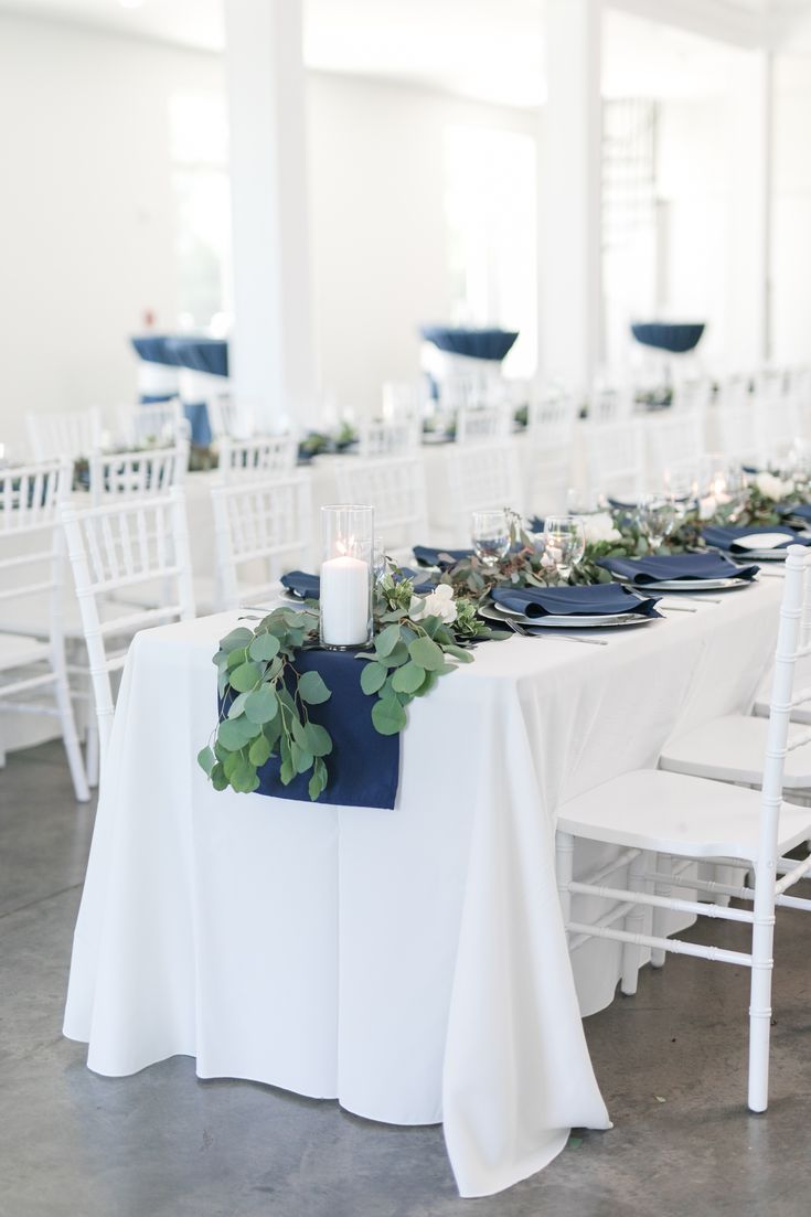 the table is set up with white chairs and blue napkins, greenery and candles