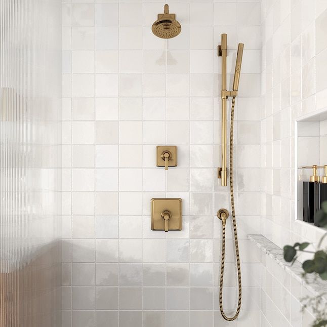 a bathroom with white tile and gold fixtures on the shower head, hand held by a brass faucet