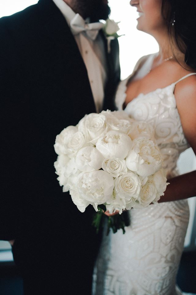 a bride and groom pose for a wedding photo in front of a window with white flowers