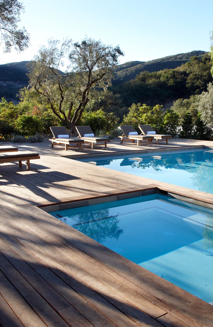 a wooden deck next to a swimming pool with chaise lounges and trees in the background