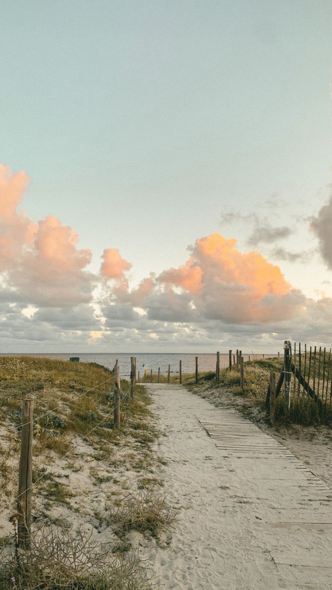 an empty path leading to the beach with clouds in the sky and grass on either side