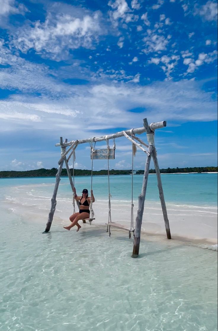 a woman swinging on a swing set in the water at an empty beach with blue skies and white clouds