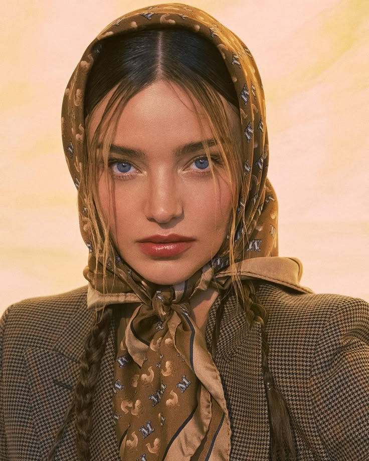 a woman with braids wearing a brown jacket and neck scarf over her head is looking at the camera