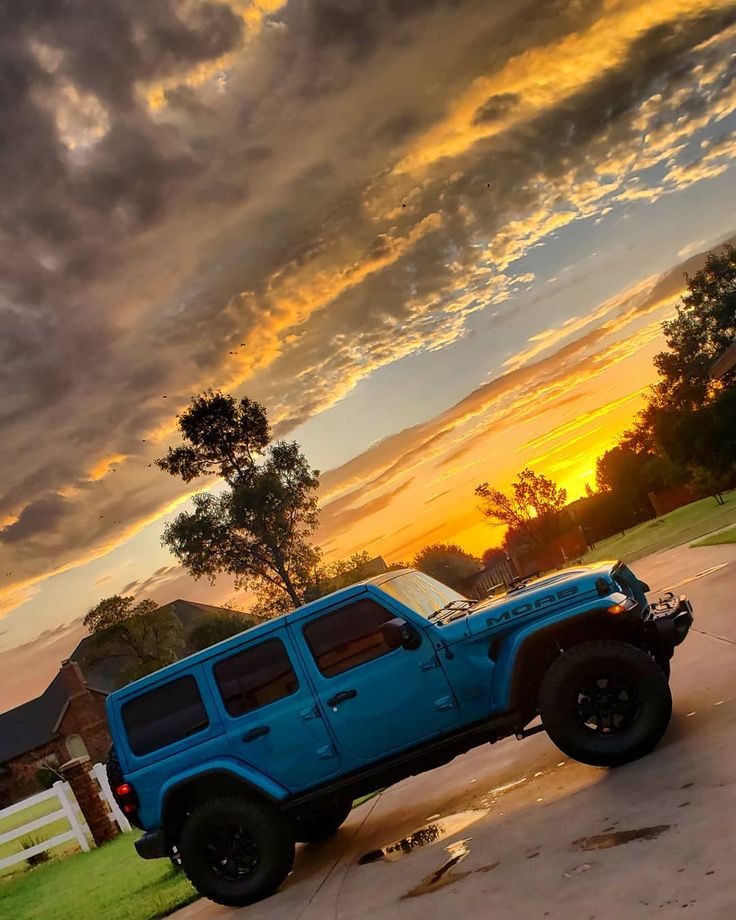 a blue jeep parked on the side of a road at sunset with clouds in the sky