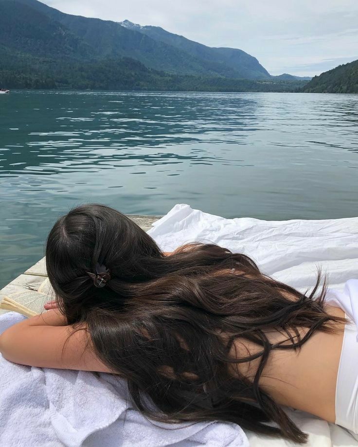 a woman laying on top of a white towel next to water with boats in the background