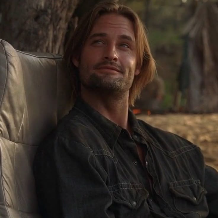 a man with long hair sitting on a couch next to a tree and looking off into the distance