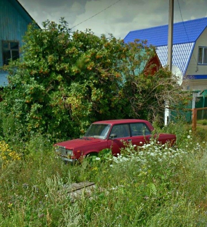 an old red car sitting in the middle of some tall grass and weeds with houses in the background