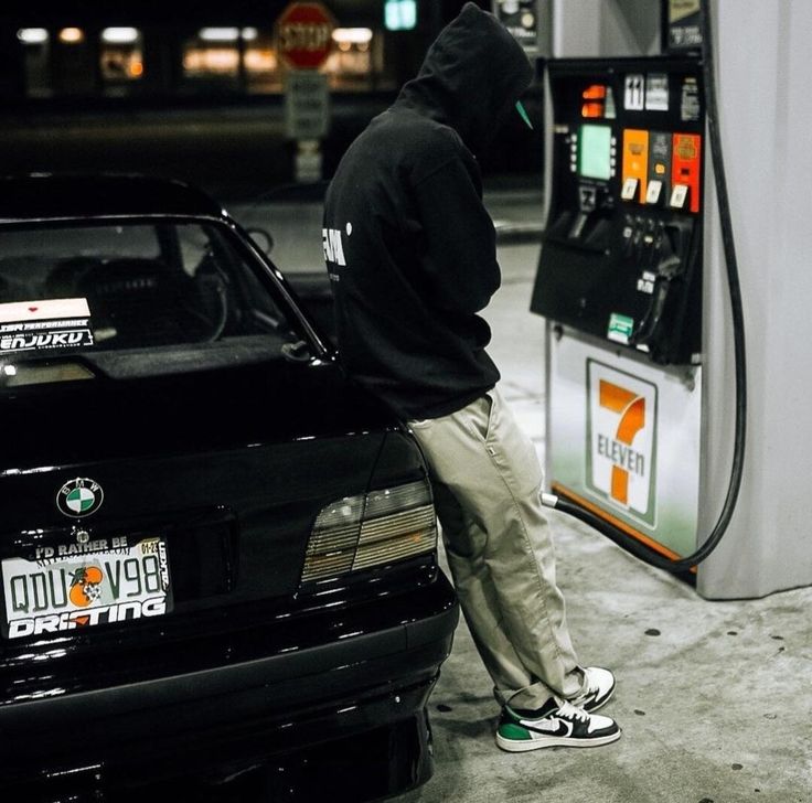 a person in a hoodie is standing next to a car at a gas pump