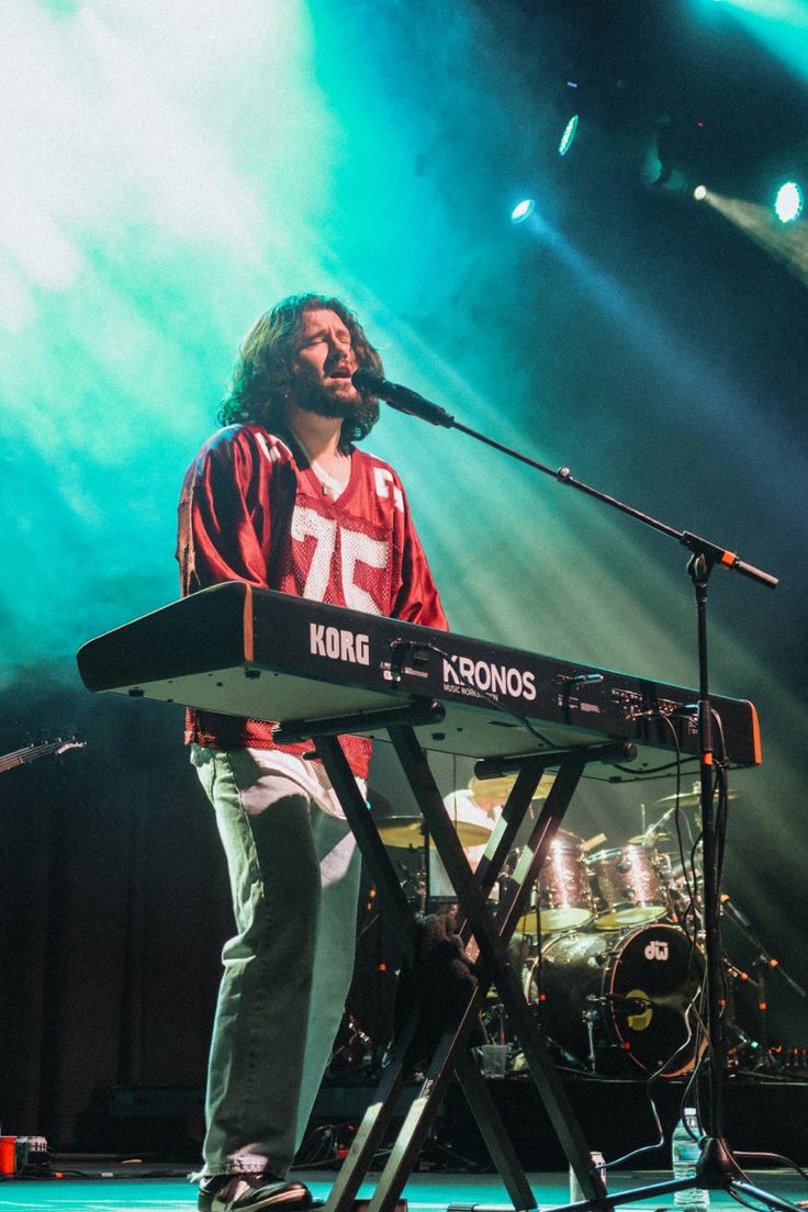 a man standing at a keyboard in front of a microphone on stage with lights behind him