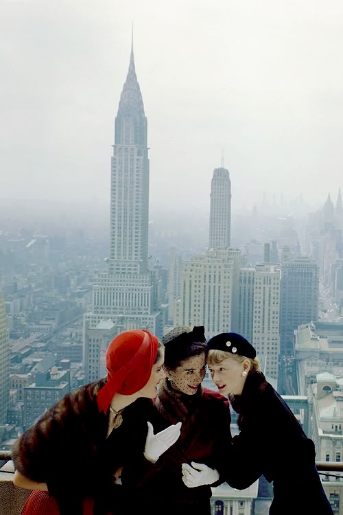 two women standing next to each other on top of a tall building in the city