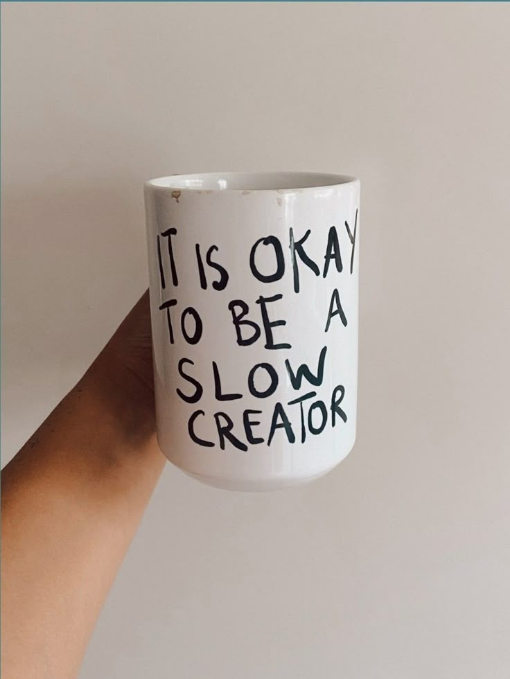 a hand holding a coffee mug with writing on it that says it is okay to be a slow creator