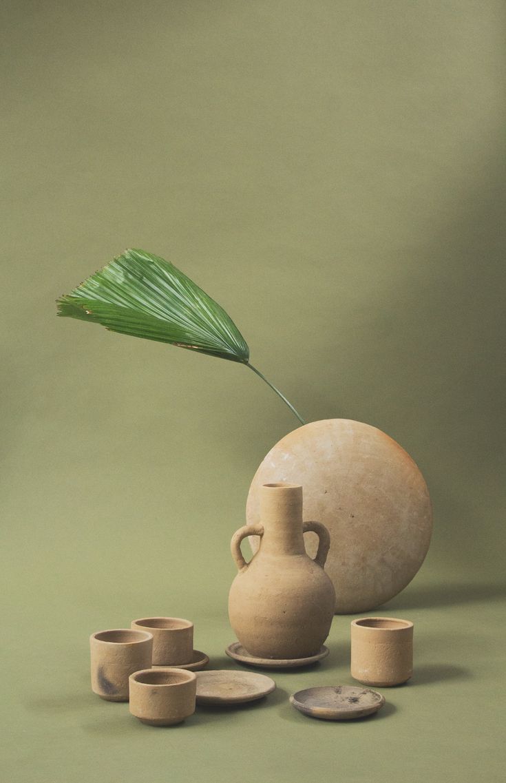 a vase with a green leaf sticking out of it, surrounded by small cups and saucers