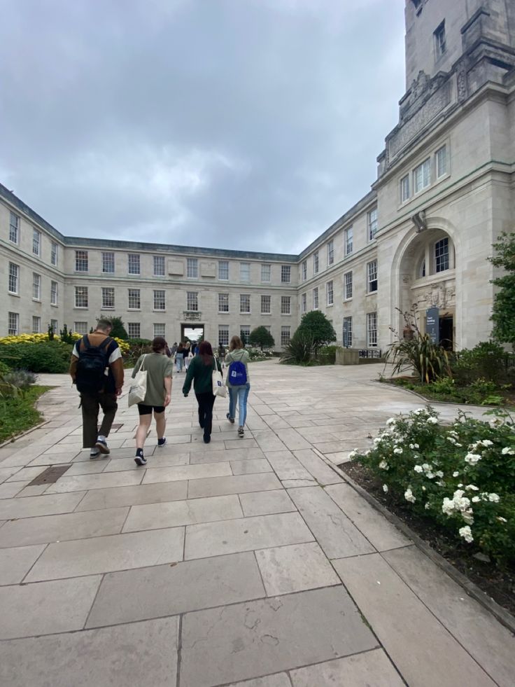 several people walking down a walkway in front of a large building with flowers on the ground