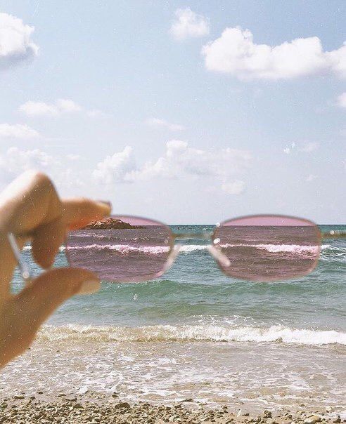 a person holding up their glasses to the camera while standing in front of the ocean