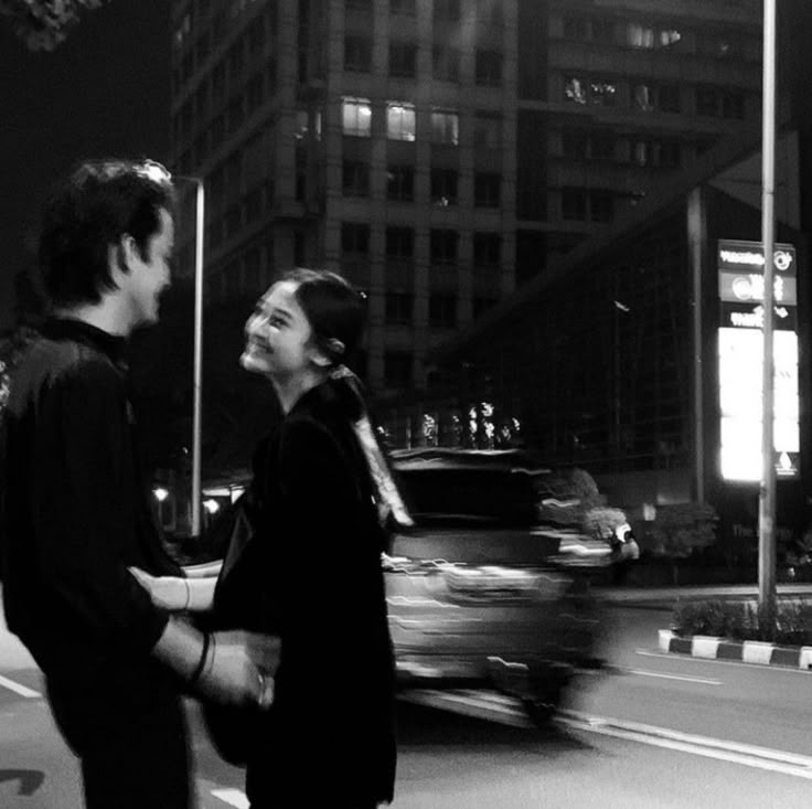 black and white photograph of two people standing in the street at night with cars passing by