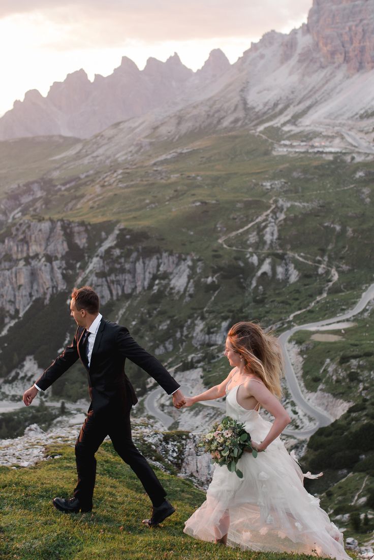 a bride and groom holding hands while walking on the side of a mountain with mountains in the background