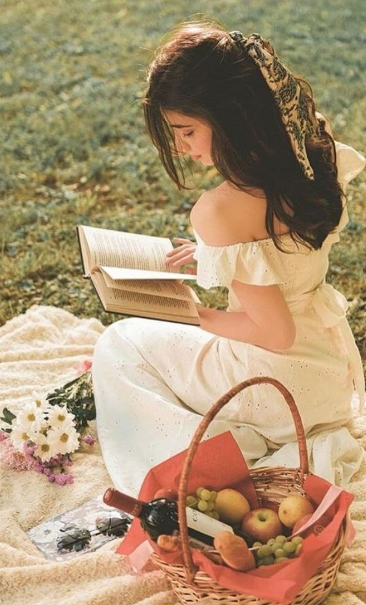 a woman sitting on a blanket reading a book next to a basket full of fruit