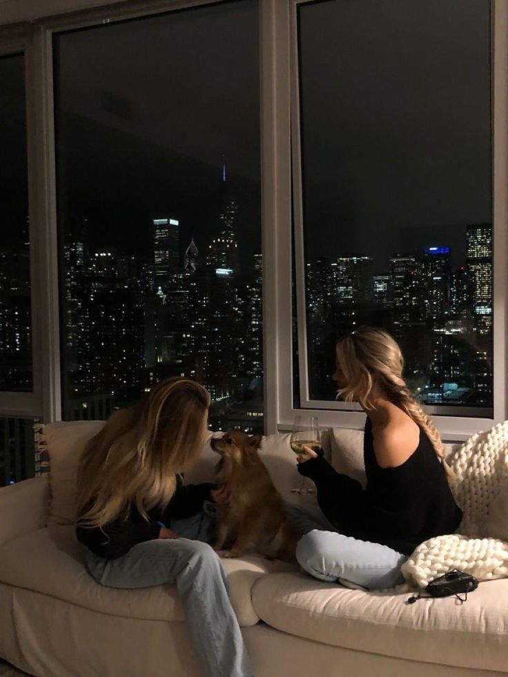 two women sitting on a couch in front of large windows with city lights behind them