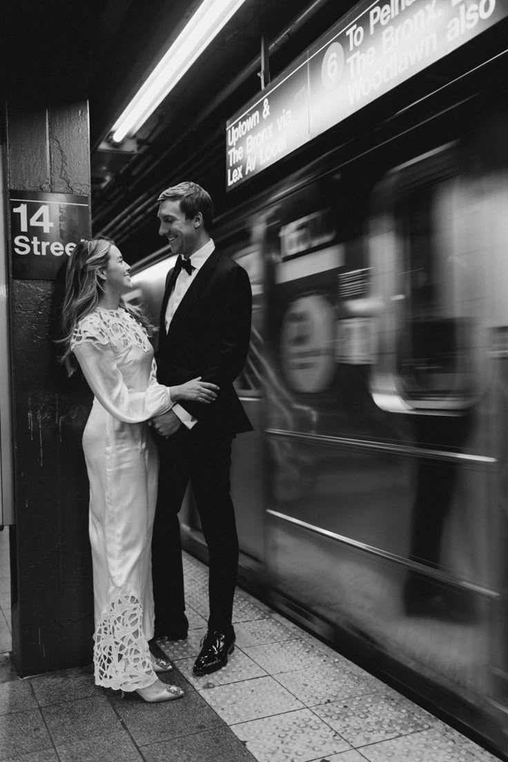 a man and woman standing next to each other on a subway platform with a train in the background