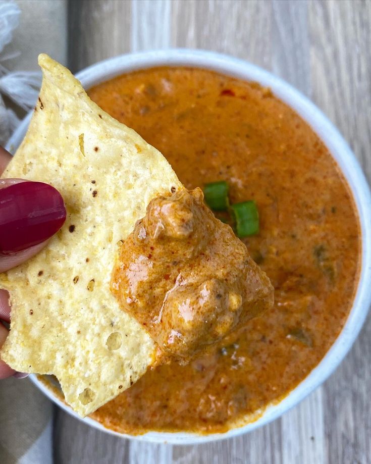 a hand holding a tortilla chip over a bowl of chili and cheese soup