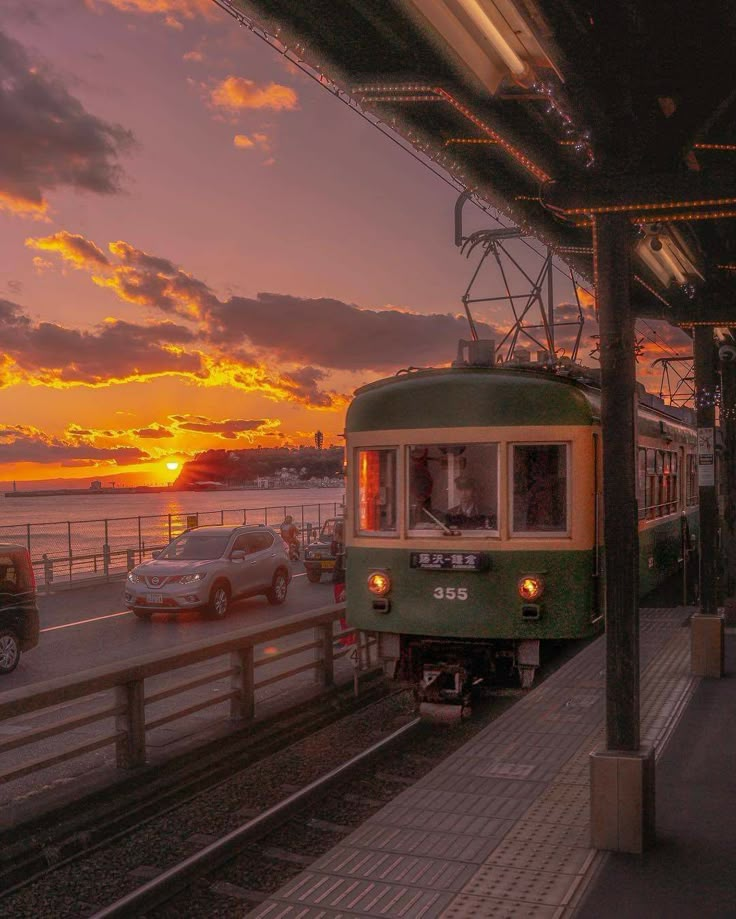 a green train traveling down tracks next to the ocean at sunset with cars passing by