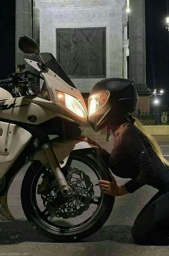 a woman kneeling down next to a motorcycle on the street at night with her headlight on