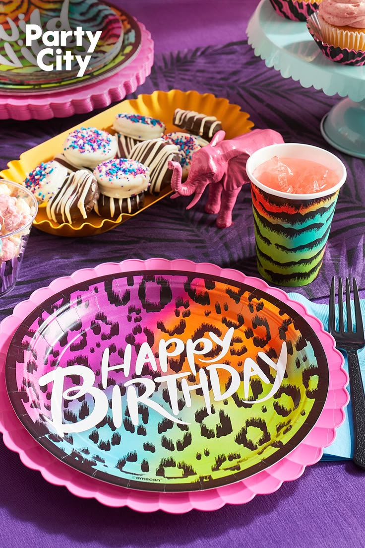 a birthday party table set up with colorful plates and cupcakes on purple cloth