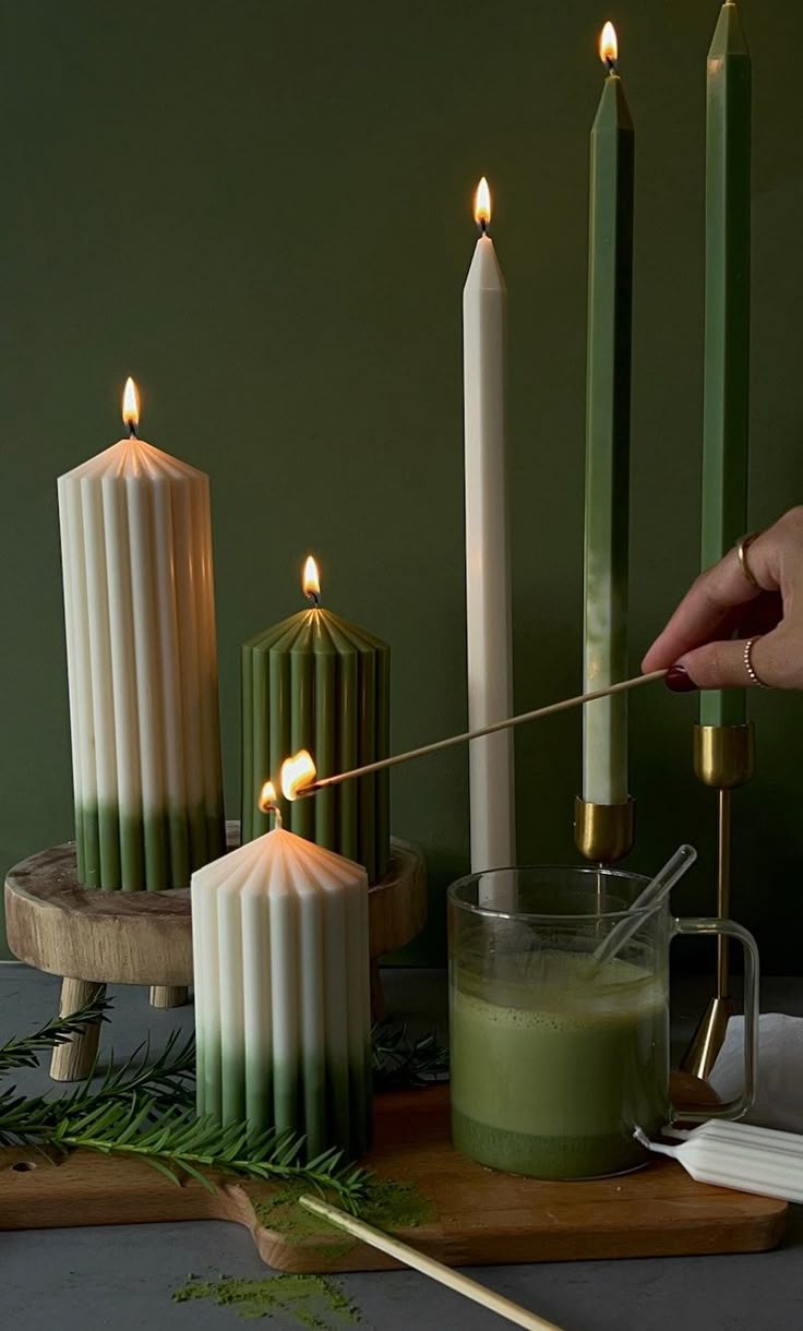 a person lighting candles on a table with green liquid and greenery next to it