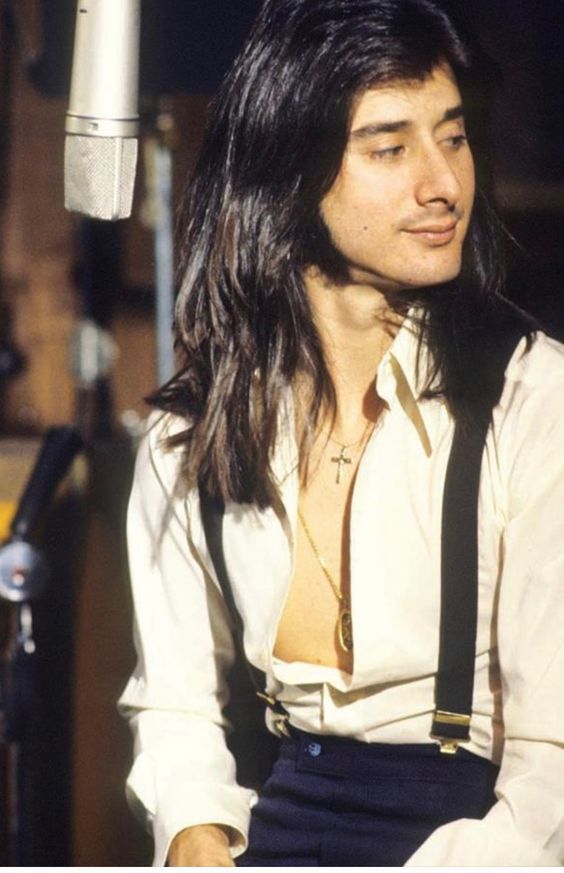 a man with long hair and suspenders sitting in front of a microphone