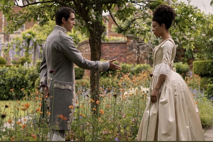 a man and woman dressed in period clothing standing next to each other on a path surrounded by flowers