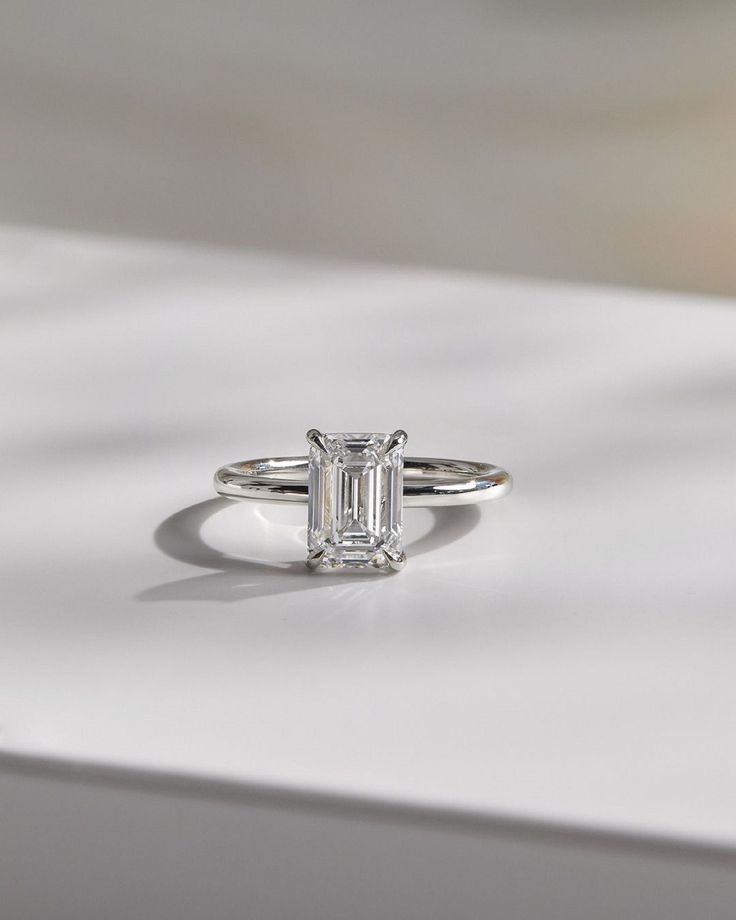 an emerald cut diamond ring sitting on top of a white surface with shadow from the background