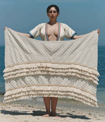 a woman standing on top of a sandy beach holding a white blanket over her face