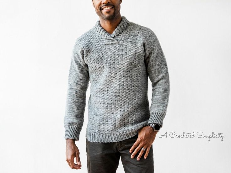 a man wearing a gray sweater and black pants standing in front of a white wall