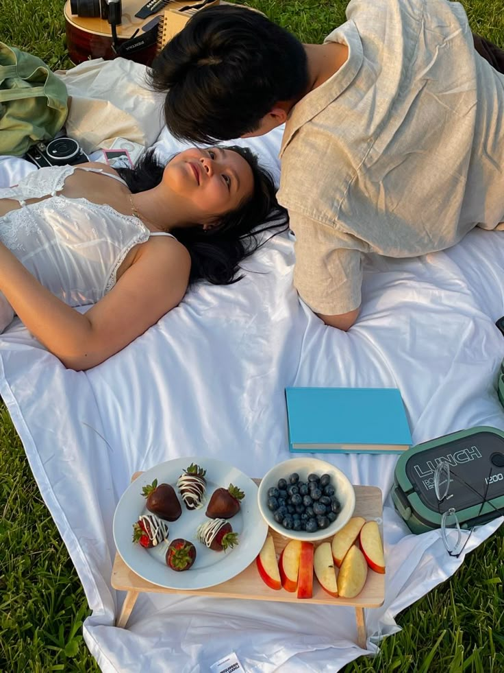 two people laying on a blanket in the grass with some fruit and chocolate covered strawberries