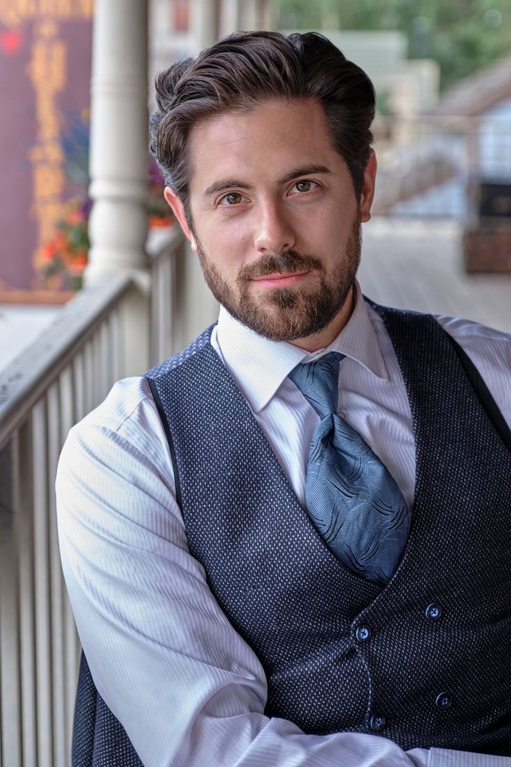 a man wearing a vest and tie standing on a porch with his arms crossed looking at the camera