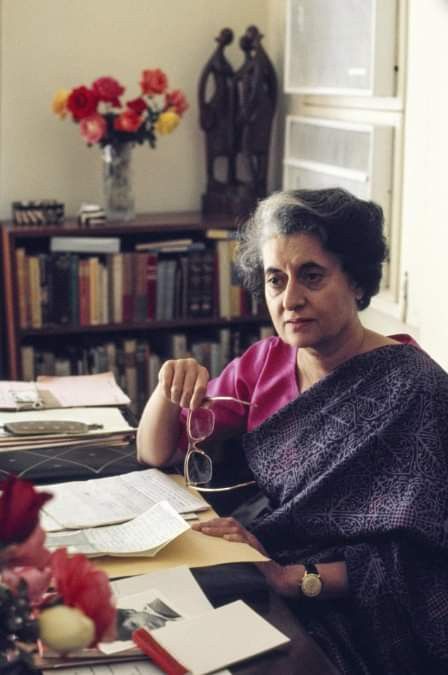 an older woman sitting at a table with papers and scissors in her hand, looking off to the side